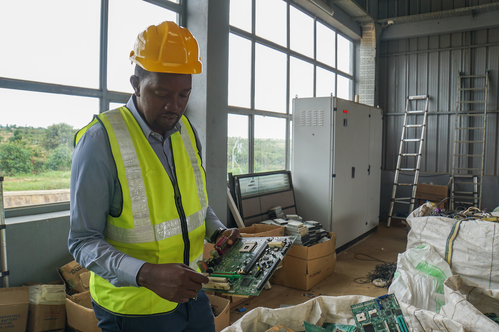 A man in a neon green vest and hard hat examines a circuit board at the Enviroserve Rwanda recycling center