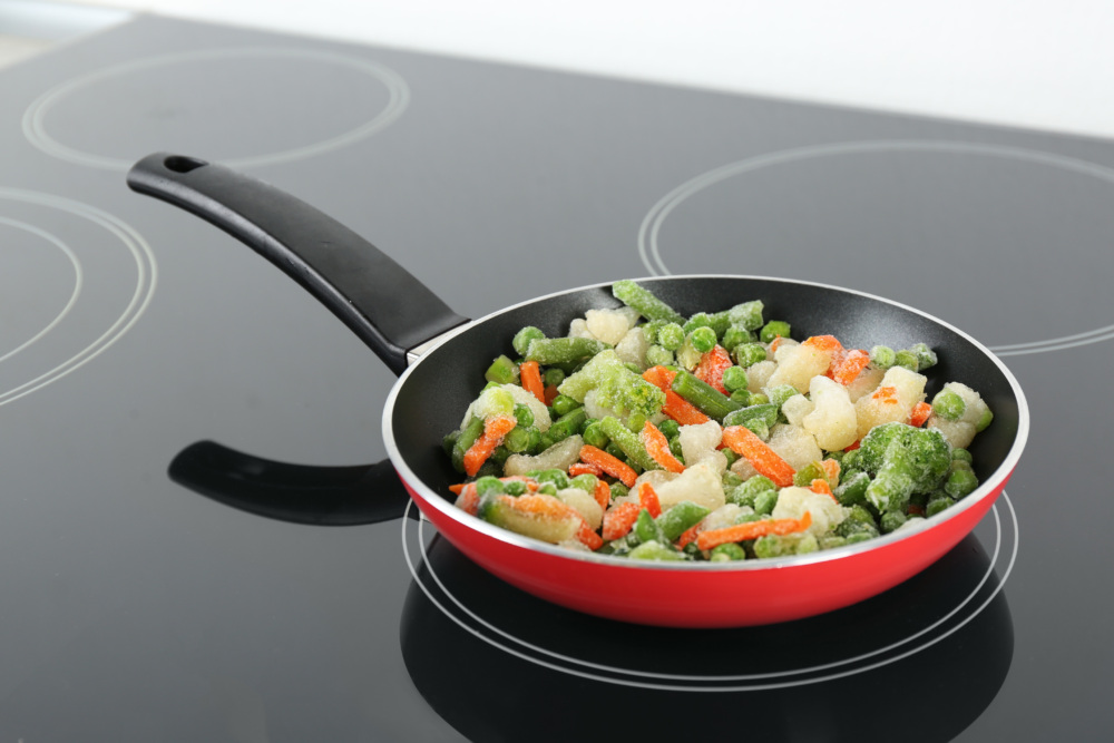 A red-bottomed nonstick pan on an induction cookstove with vegetables cooking.
