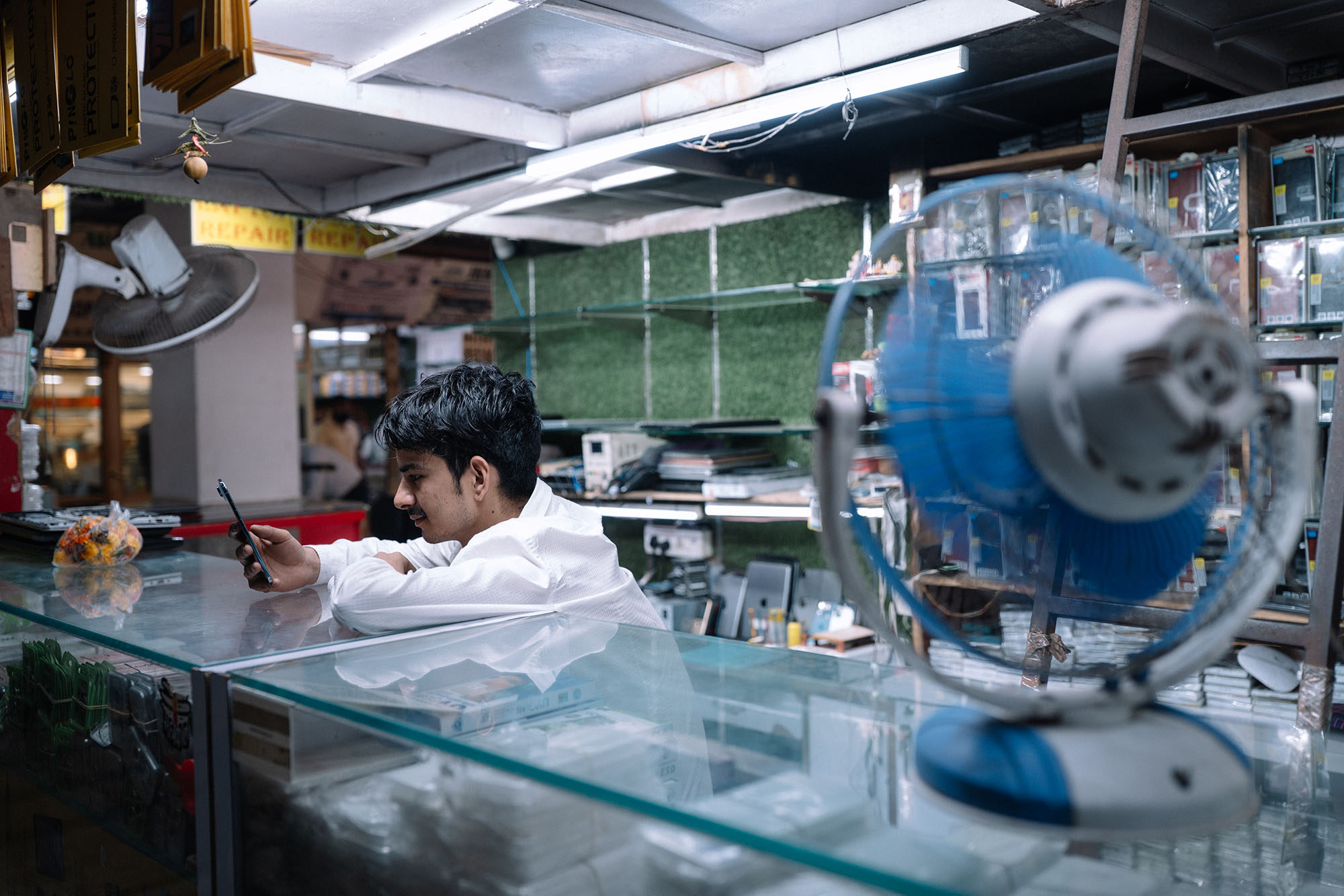 A young man standing behind a counter looks at his phone and smiles while a fan blows air on him.