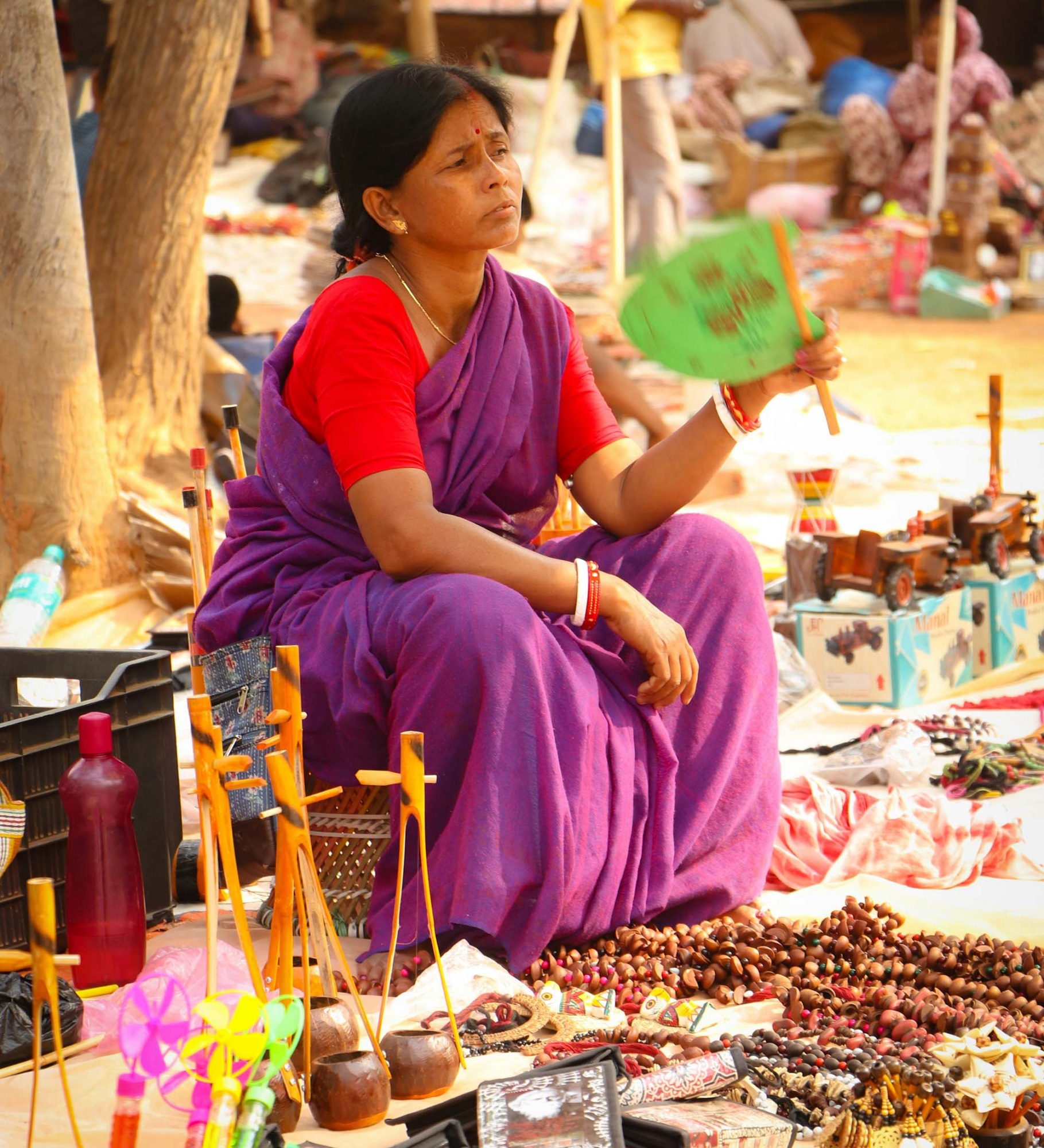A woman wearing a red and pink sari sits and fans herself with a green fan while selling handicrafts.