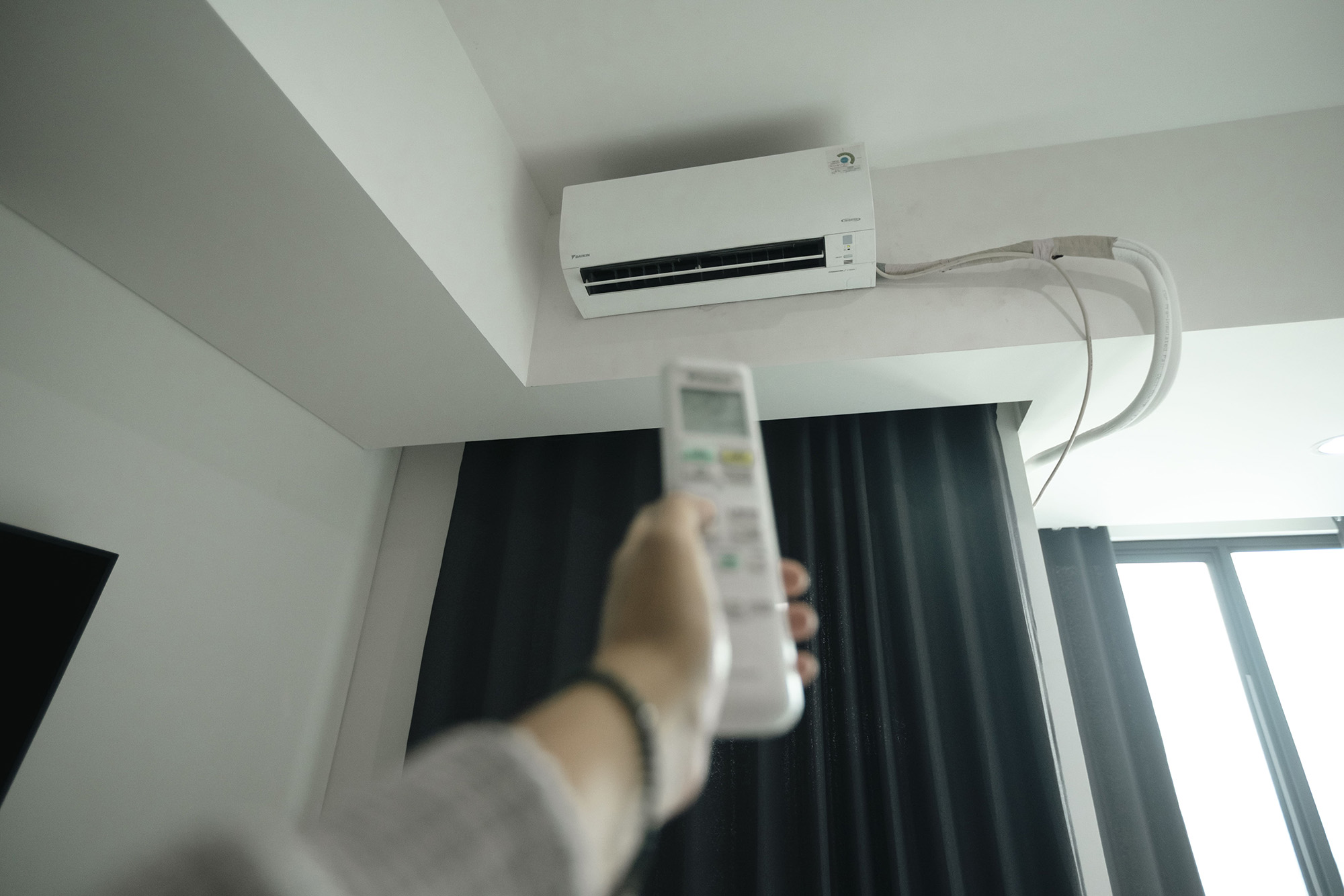 A hand holding a remote control points to a split air conditioning unit.