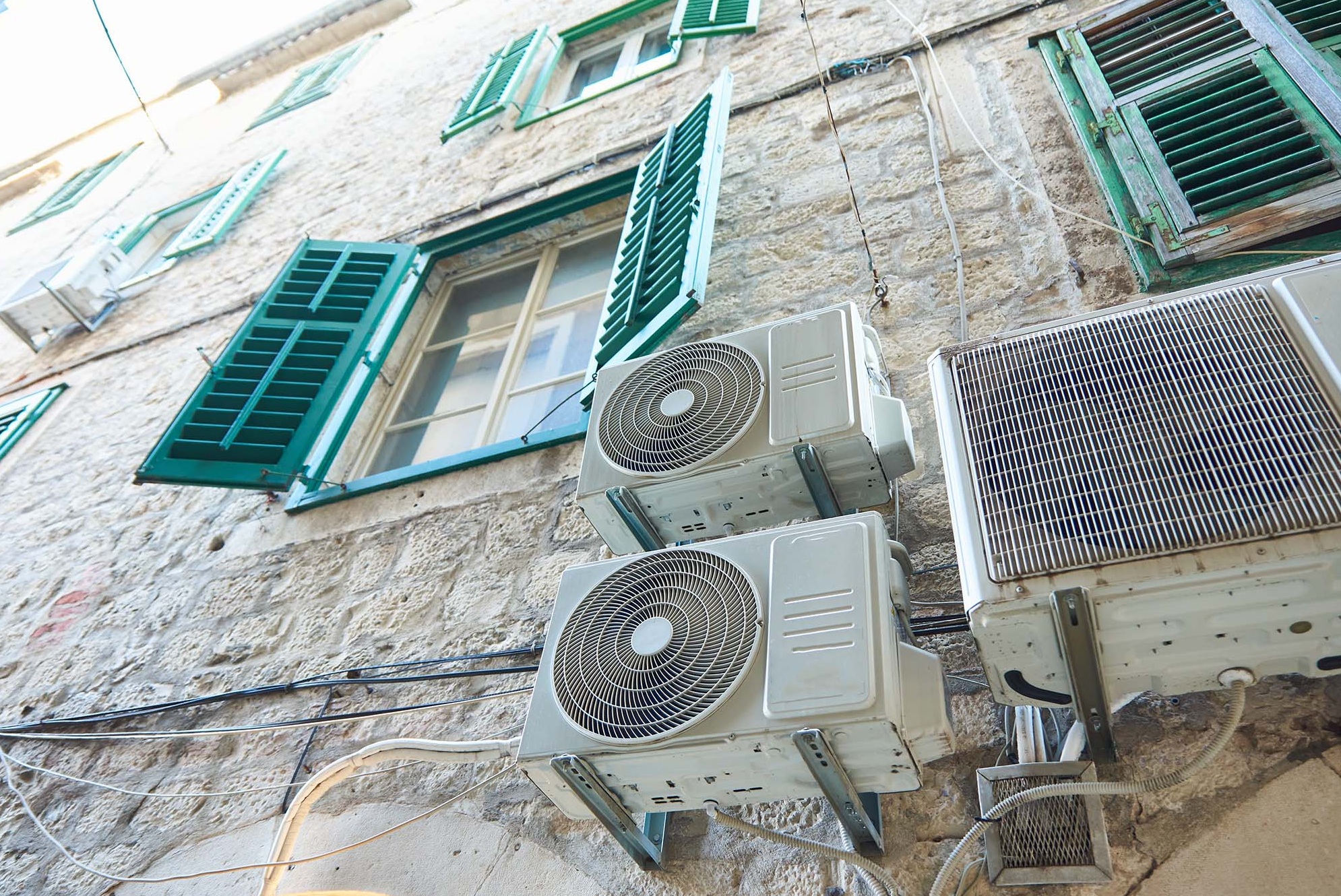 Air conditioners attached to the exterior of a building whose windows have green shutters